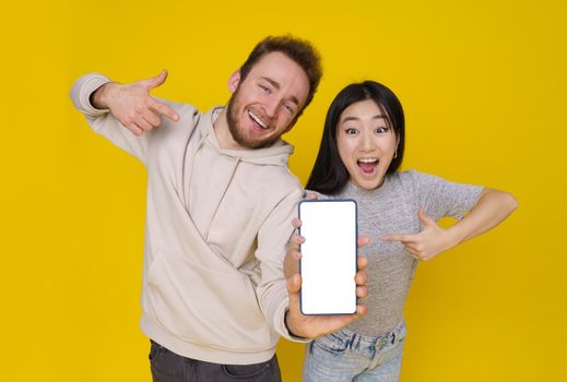 Caucasian guy and asian girl show thumbs up excited holding smartphone with blank white screen, mobile app advertisement isolated on yellow background. Product placement. Great offer.