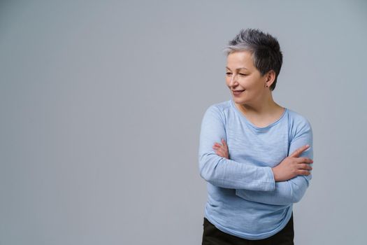 Happy mature grey hair woman posing with hands folded looking sideways up, copy space on left isolated on white background. Healthcare concept. Aged beauty concept.