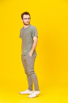 Young man wears eye glasses in casual wear posing on yellow background with hands in pockets looking sideways. Stylish bearded smart casual look hipster man. Thoughtful guy on yellow background.