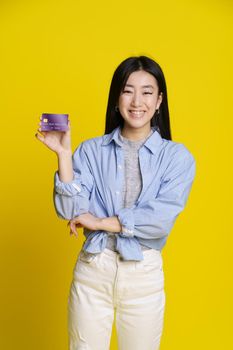 Smiling asian girl holding credit, debit card in hand. Asian girl in blue shirt with mockup banking card isolated on yellow background. E-banking concept. E-commerce concept.