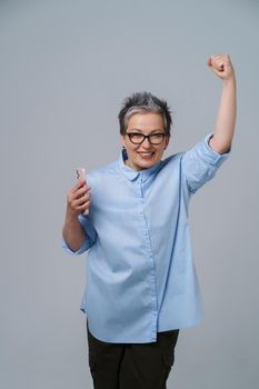 Mature business woman with phone in hand raised one hand up celebrating victory, success. Pretty woman in blue shirt isolated on white background.