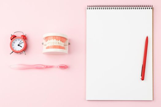 Open spiral notepad with pen and alarm clock on pink background. Top view. Artificial jaw and toothbrush on the dentist table.