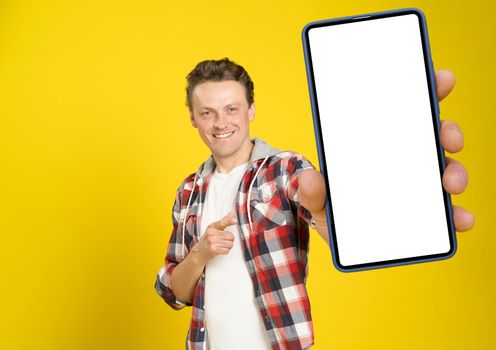 Happy to win handsome man pointing at smartphone with white empty screen, wearing red plaid shirt and jeans cellphone display mock up isolated on yellow background. Mobile app advertisement.