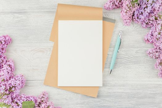 Open craft notebook with pen and lilac flowers on a white wooden background. Top view.