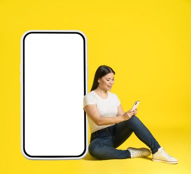 Mature asian woman leaned on huge, giant smartphone sitting on floor with phone in hands in casual white screen, looking at phone mobile app advertisement isolated on yellow. Product placement.