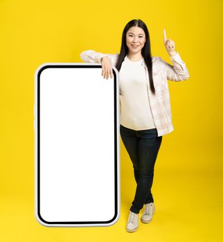 Asian woman pointing finger one as attention leaned back on giant, huge smartphone with blank white screen, wearing casual isolated on yellow background. Free space mock up.