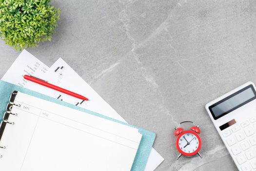 Spiral notebook in soft blue cover with a metal pen, calculator, alarm clock, stock charts and a flower in a pot on a gray marble background. Office desk concept. Top view.