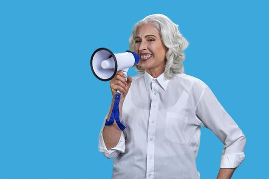 Expressive senior woman in white blouse talking into megaphone. Mature lady with loudspeaker on blue background.