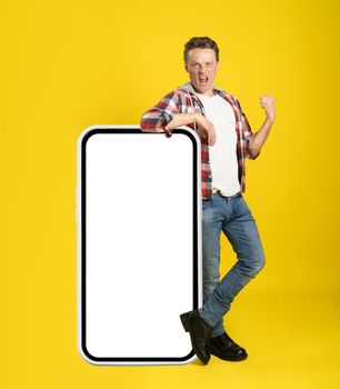 Pointing on huge, giant smartphone with white screen young man leaned on it gesturing with another hand dressed in casual isolated on yellow background. Free space mock up.