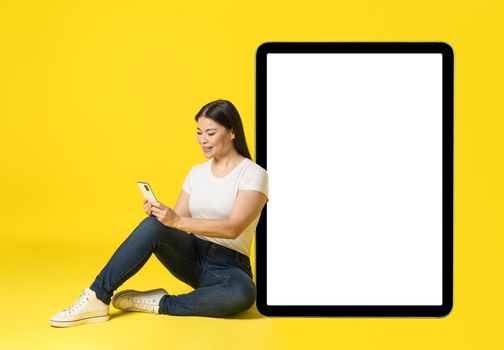 Asian woman leaned on huge, giant tablet pc sitting on floor with phone in hands in casual white screen, looking at phone mobile app advertisement isolated on yellow. Product placement.