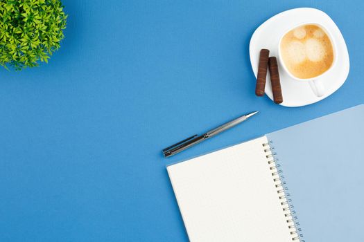 Spiral open notebook with a metal pen, dessert chocolate sweets, cup of coffee and home plant with green leaves on blue background. Flat lay, top view.