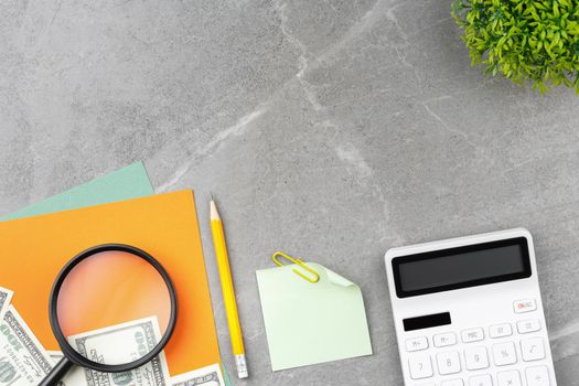 Spiral notebook, pencil, stock charts, calculator, magnifying glass, note paper and a flower in a pot on a gray marble background. Top view. Office desk concept.