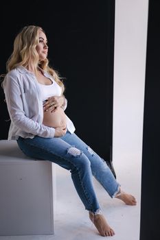 Pregnant woman sits on white cube an put hands on belly. Denim style, white shirt and jeans. Blond hair lady barefoot.