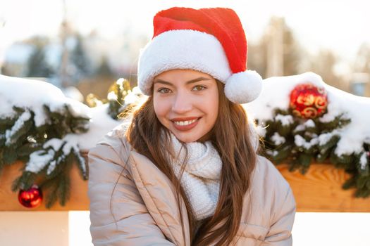 Winter portrait young adult beautiful woman in Santa hat. Christmas mood. Snowing. Winter beauty fashion concept. Pretty young woman sitting bench outdoor near Christmas garland