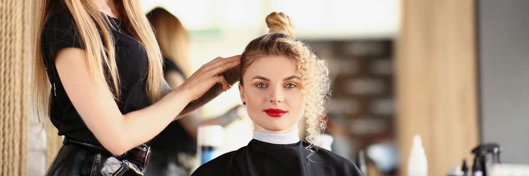 Portrait of professional female stylist creating hairstyle for client doing wavy hair. Blonde lady in hairdresser seat get ready for party. Beauty concept