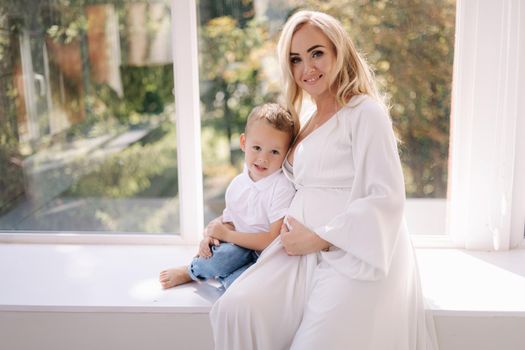 Beautiful pregnant woman with her son. Boy hug his mom and rejoices. Boy waiting for sister. Lady in elegant white dress posing to photographer in studio. Background of white tulle.