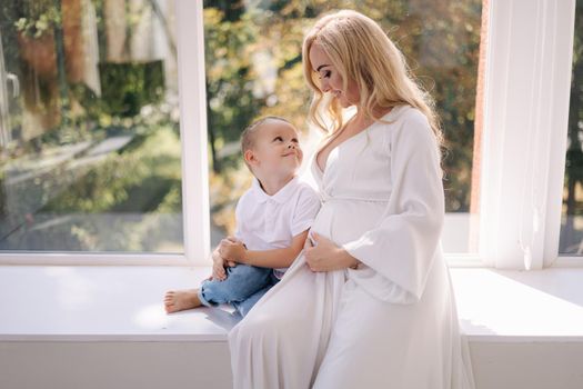 Beautiful pregnant woman with her son. Boy hug his mom and rejoices. Boy waiting for sister. Lady in elegant white dress posing to photographer in studio. Background of white tulle.