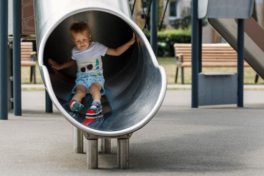 Boy in a white t-shirt blonde, riding on a metal slide tube on the playground.