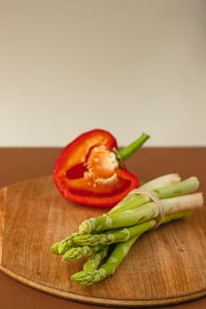 branches of fresh green asparagus on a wooden board, half of a red big pepper in the background. brown and light beige background. Basic trend concept with copy space