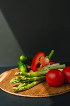 Vegetables lie on a wooden board: tomatoes, asparagus, cucumbers, red bell peppers. brown, dark gray background. place for text