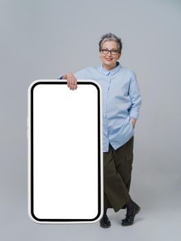 Grey hair mature business woman leaned on giant, huge smartphone with white, black screen wearing casual blue shirt isolated on white background. Free space mock up.