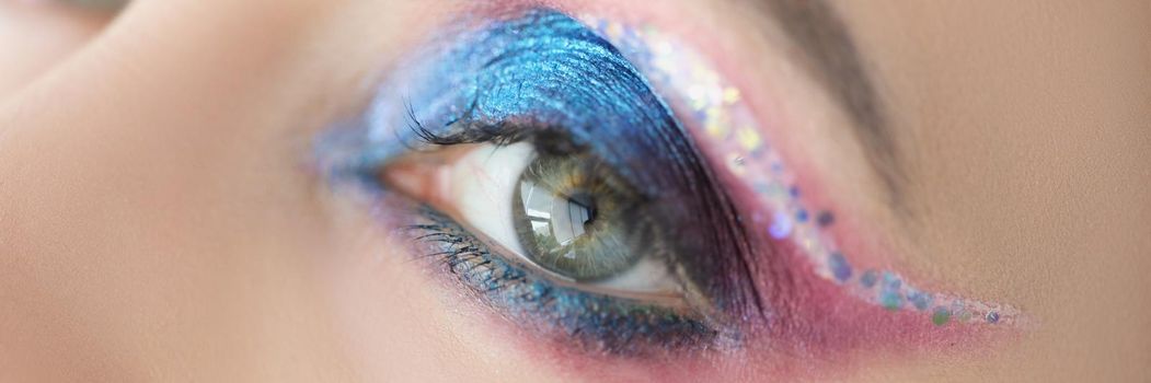 Close-up of festive bright eye makeup on womans face with blue and pink glitter. Cats eye technique and colourful mix of eyeshadows. Mua, creative concept