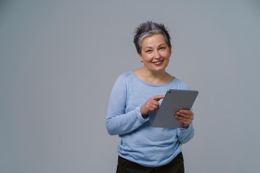 Mature grey haired businesswoman with digital tablet in hands working online. Pretty woman in 50s in blue blouse isolated on white. Older people and technologies.