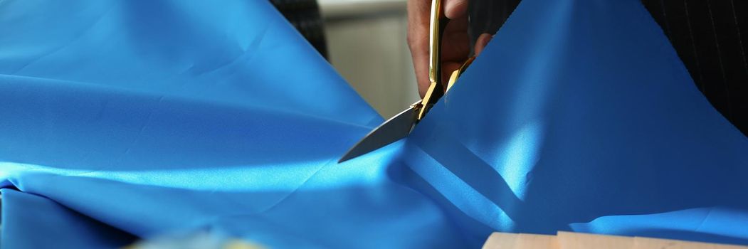 Close-up of atelier worker cutting blue cloth with scissors for further sewing. Yellow thread on table, creative process at cloth studio. Fashion concept