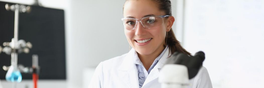 Portrait of pretty cheerful laboratory worker in medical gown examine sample under microscope. Smart chemist and flasks on desk. Science, explore concept