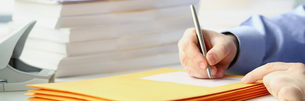 Close-up of man with pen in hand signing envelope while sitting at table with bunch of documents. Prepare documents to send to recipients. Mail concept