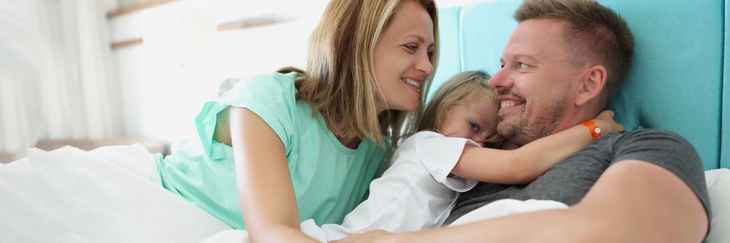 Portrait of happy cheerful whole family hugging in bed together in morning. Smiling father mother and little girl on weekend. Family, relationship concept