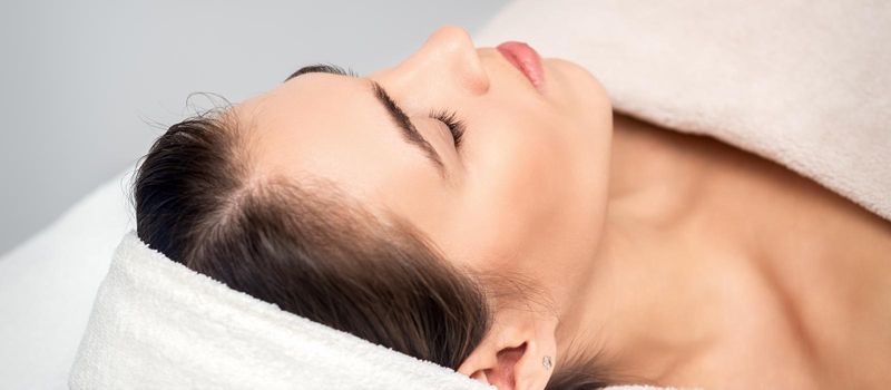 Side view of young woman lying on beautician table with closed eyes while waiting for cosmetic procedure in beauty salon