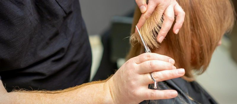 Back view of hairdresser cuts red or brown hair to young woman in beauty salon. Haircut in hair salon. Soft focus