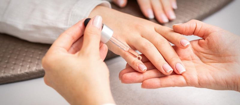 Hand of manicurist pours oil by pipette to cuticle of nails of young woman in beauty salon. French manicure