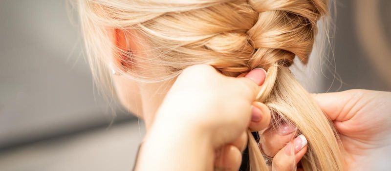 Close up back view of female hands braiding a pigtail to young blond woman at beauty salon