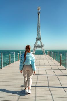 Large model of the Eiffel Tower on the beach. A woman walks along the pier towards the tower, wearing a blue jacket and white jeans