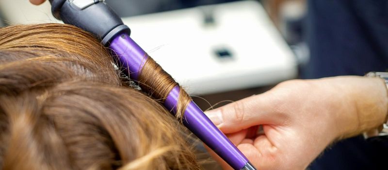 Professional hairdresser makes curly hairstyle by curling iron for long red hair of young woman in hair salon