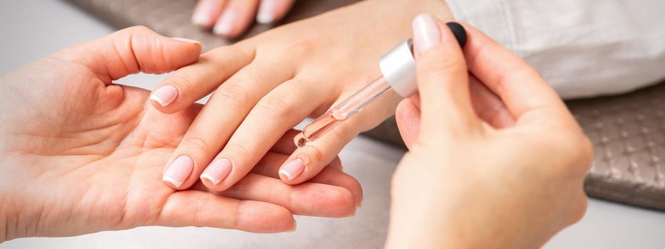 Hand of manicurist pours oil by pipette to cuticle of nails of young woman in beauty salon. French manicure