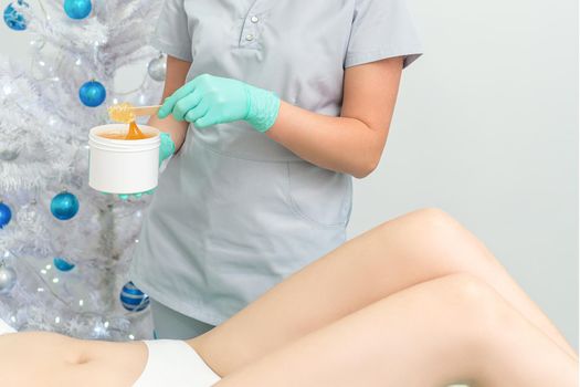 Female cosmetologist holding white blank jar with sugaring paste on christmas tree background