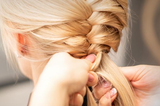 Close up back view of female hands braiding a pigtail to young blond woman at beauty salon