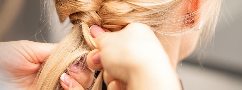 Close up back view of female hands braiding a pigtail to young blond woman at beauty salon