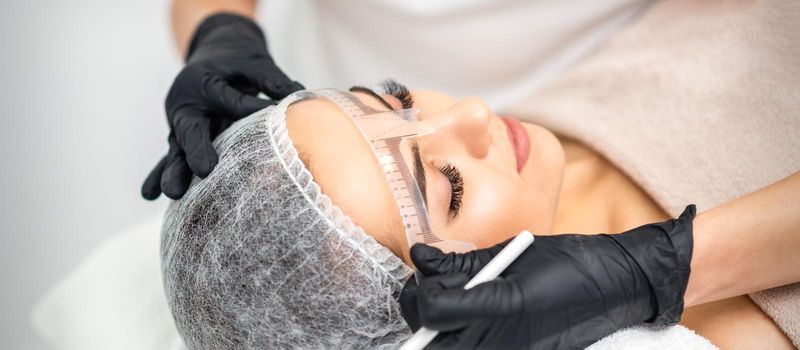 Cosmetologist is measuring with ruler the brows of young caucasian woman before permanent makeup tattoo