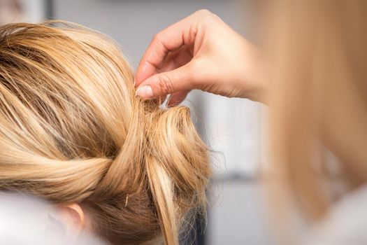 Close up back view of hand of female hairdresser makes hairstyle of young blonde woman in beauty salon