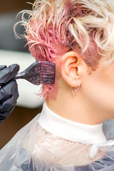 Hand of hairdresser dyeing hair in pink color by paintbrush on back of head of young caucasian woman in beauty salon. Selective focus