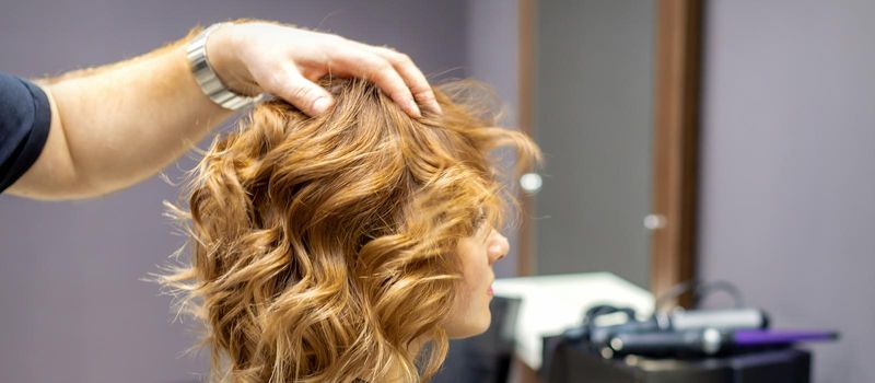 Side view of male hairdresser checks curly hairstyle of young caucasian woman in hair salon