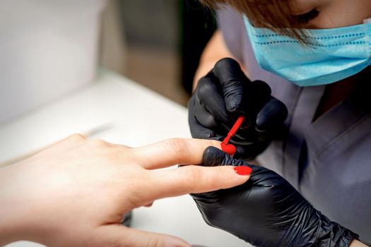 Manicurist with protective mask and gloves applying red nail polish to female nails in nail salon