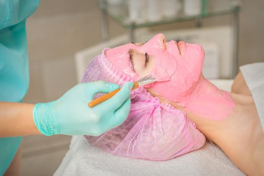 Hand of cosmetologist applying pink alginic mask to face of young woman in beauty salon