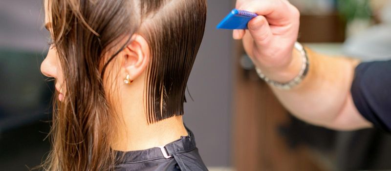 Side view of hand of hairdresser doing haircut of young woman by comb in hair salon