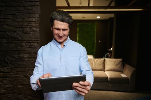 Pleasant handsome Caucasian mature grey-haired businessman, entrepreneur, interior designer planning a new design project, working on a digital tablet in a cozy stylish interior lit with dim light