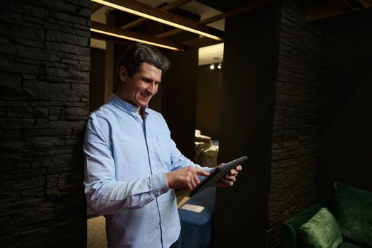 Confident prosperous sales man, real estate agent, interior designer working at a furniture store, using digital tablet and checking orders of new upholstered furniture in an interior design showroom